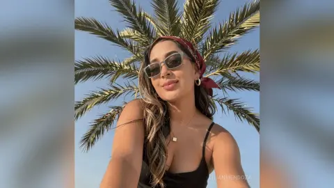LilianMendoza poses outdoors against a palm tree, wearing sunglasses, a red headband, and a black tank top.