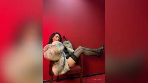 Glamorous woman in a luxury faux fur coat and black lingerie relaxing on a chair against a red wall.
