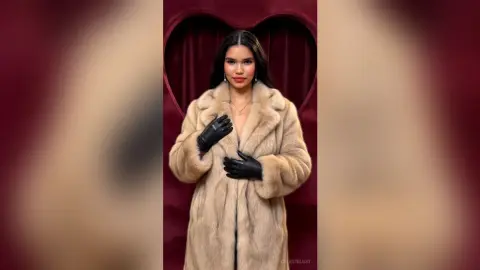 Elegant woman in a luxurious beige faux fur coat posing against a romantic red heart-shaped velvet backdrop.
