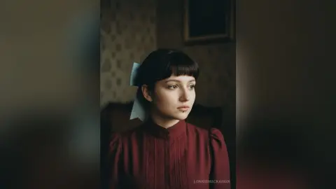 Melancholic young woman with dark bangs and white hair bow wearing a vintage burgundy high-neck blouse.