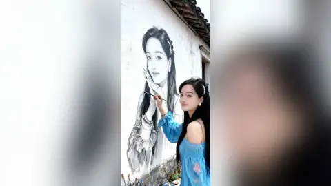 Young woman in a blue dress painting a large black and white portrait mural on an outdoor building wall.