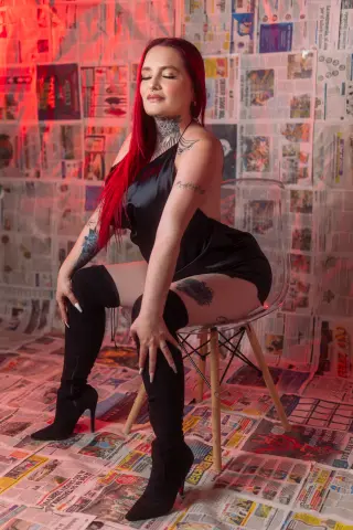 AlejaMartinezz with long red hair sits on a clear chair wearing a black slip dress and tall black boots against a wall of newspapers.