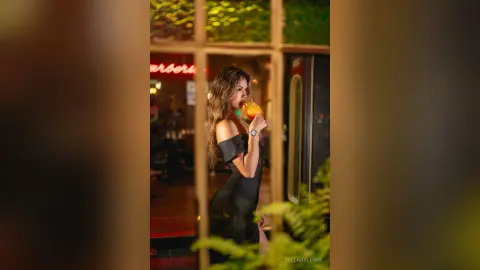 Stylish woman in a black off-the-shoulder dress sipping a cocktail at a moody nighttime outdoor cafe.