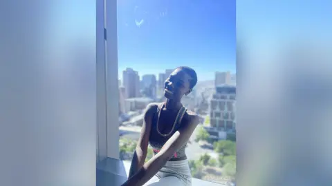 Smiling young woman in a grey tank top relaxing by a bright window overlooking a sunny city skyline.