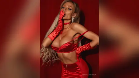 LeslyAnderson poses in red satin lingerie and matching gloves, showing off her cleavage against a red background.