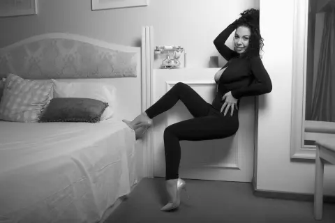 Smiling woman in a black bodysuit posing by a bed in a bright, minimalist bedroom setting.