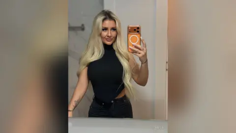 Anita poses for a mirror selfie wearing a black sleeveless turtleneck and black jeans, showing off her long blonde hair and arm tattoos.