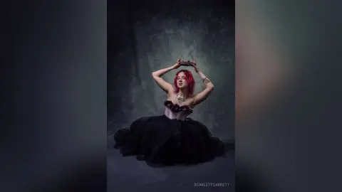 ScarlettGarrett wears a black tulle gown and holds a gold headpiece above her pink hair against a dark grey background.