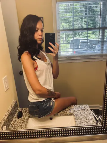 Young woman in a white tank top and denim shorts taking a mirror selfie in a bright indoor setting.