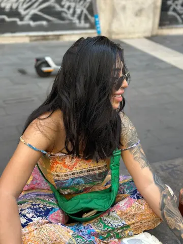 Smiling woman in colorful floral print outfit sitting on a city street with long dark hair and sunglasses.