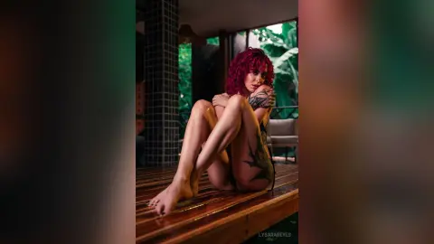 Red-haired woman with tattoos sitting on a wooden deck in a lush, moody tropical garden setting.