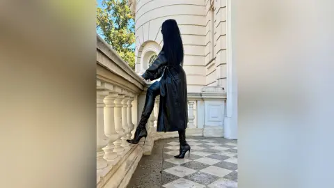 Stylish woman in a black leather trench coat and high heels posing on a classic stone balcony.