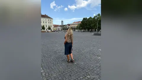 SelenaRusse stands in a cobblestone city square wearing a dark blue dress and sandals, looking away from the camera.
