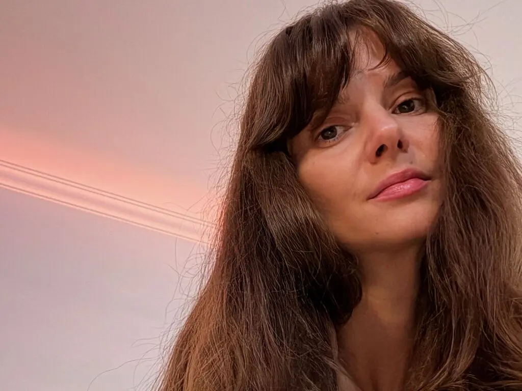 Close-up portrait of a brunette woman with long hair looking thoughtfully at the camera in warm indoor lighting.
