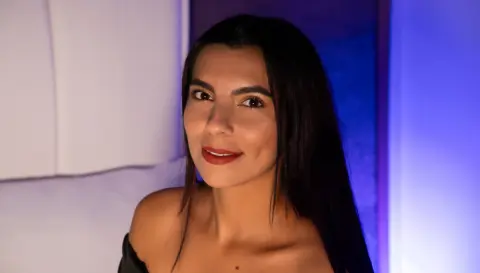 Smiling young woman with long dark hair in a black off-the-shoulder top against a moody blue lit background.