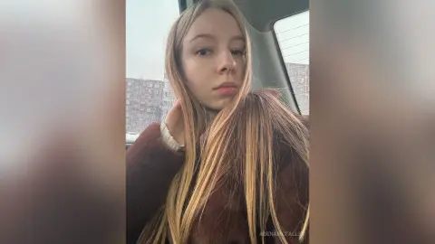 AdenaMcfalls poses in a brown furry coat with her long blonde hair falling over her shoulder while sitting in a car.