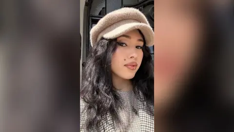 Young woman with long dark hair wearing a beige beret and grey patterned sweater in soft natural lighting.