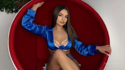 Young woman in a blue satin crop top and denim shorts relaxing in a large red velvet chair.