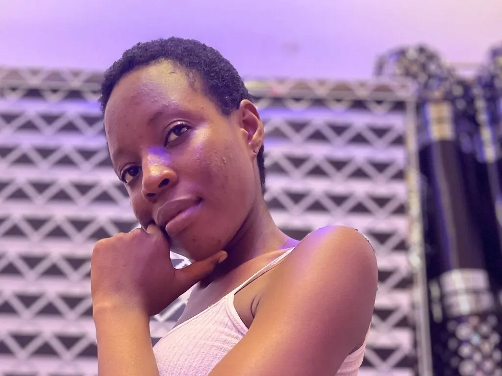 Thoughtful young woman in a white ribbed tank top posing against a patterned studio background with purple lighting.
