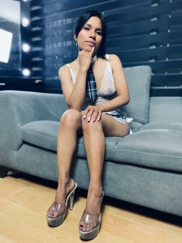 Elegant woman in a white lace dress and clear high heels sitting on a grey sofa in a modern living room.