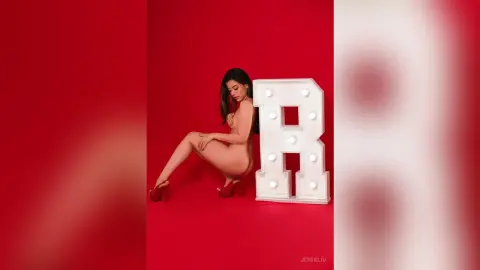 JessieLiu poses naked against a red background, sitting next to a large lit letter R while touching her thigh.