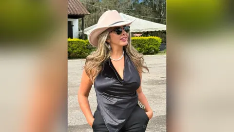 CrisstyBaez — CrisstyBaez wears a black satin halter top and a wide-brimmed hat while posing outdoors in the sun.