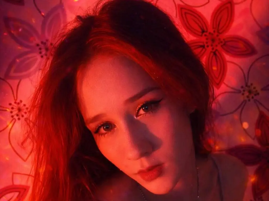 Close-up portrait of a young woman with red hair posing against a floral pattern background in warm red lighting.