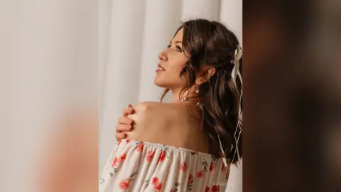 Elegant woman in a white floral off-the-shoulder blouse looking away in a soft, dreamy indoor setting.