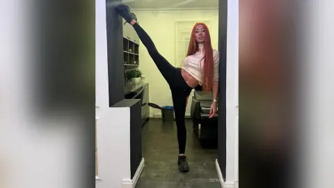 DeviNessa stands in a doorway wearing a tan crop top and black leggings, lifting one leg high against the wall.