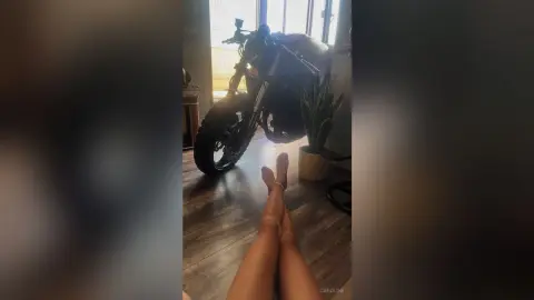 Relaxed woman resting her bare feet on a wooden floor near a motorcycle in a sunlit room.