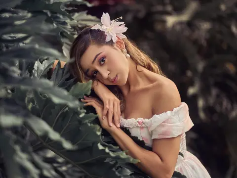 Dreamy portrait of a woman in a white floral dress leaning against lush tropical leaves in a garden setting.