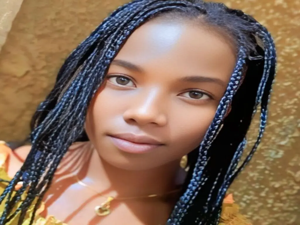 Young woman with long black braids wearing a gold necklace in a warm, sunlit indoor setting.