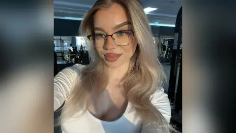 Smiling blonde woman in a white ribbed top wearing glasses in a bright gym setting.
