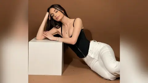 Young woman in a black tank top and white jeans leaning on a white pedestal against a brown studio background.