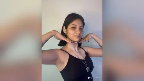 JacquiKozma poses with her hands under her chin, wearing a black tank top and a silver necklace against a plain white wall.