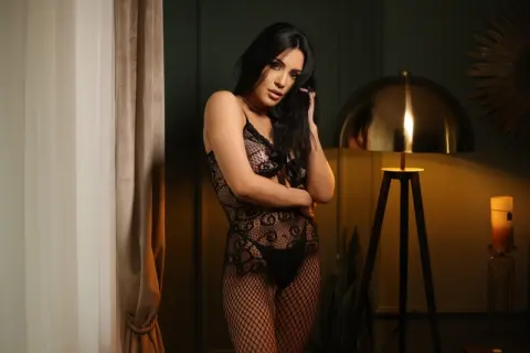 Elegant woman in black lace lingerie posing in a moody, dimly lit room with warm lamp lighting.