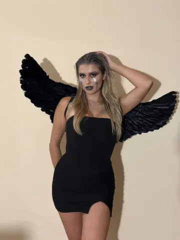 Woman in a black dress and black angel wings posing with moody makeup against a beige background.