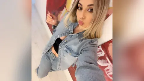 Blonde woman in a denim jacket and jeans posing playfully against an abstract painting background.