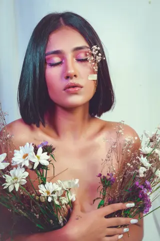Dreamy portrait of a young woman with short dark hair holding white wildflowers against a soft blue background.