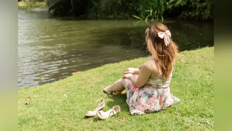 Young woman in a floral sundress sitting on a green grass bank by a peaceful lake during a sunny summer day.