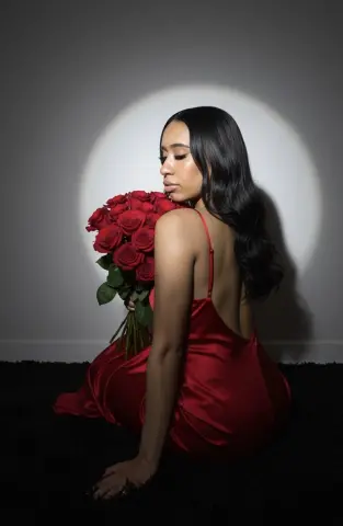 Elegant woman in a red satin dress holding a large bouquet of red roses in moody studio lighting.