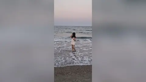 SaraTray stands in the shallow ocean waves at a beach, wearing a short white sundress while looking out toward the sea at sunset.