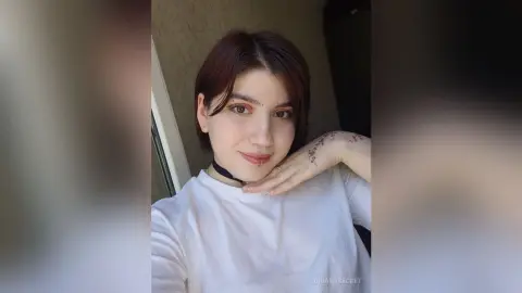 OrianaSecret — Young woman with short brown hair and a white t-shirt smiling in a soft indoor setting.