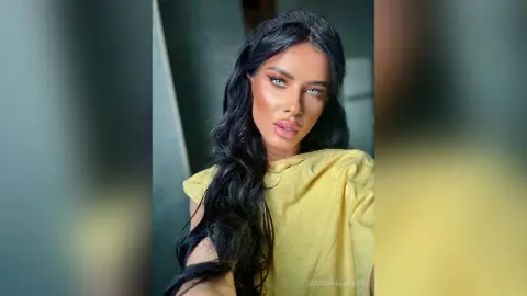 Glamorous woman with long wavy dark hair wearing a yellow top in a soft-lit indoor setting.