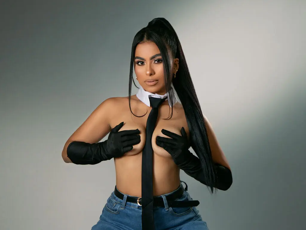 Glamorous woman in a black tie, white collar, and black gloves posing against a grey studio background.