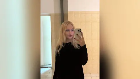 Blonde woman in a black long-sleeve shirt taking a moody mirror selfie in a tiled bathroom.