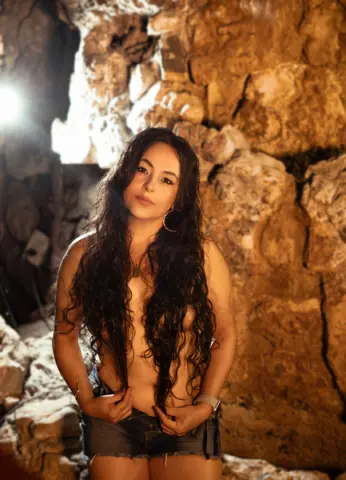 Young woman with long wavy hair in denim shorts posing in a rustic stone cave with warm lighting.