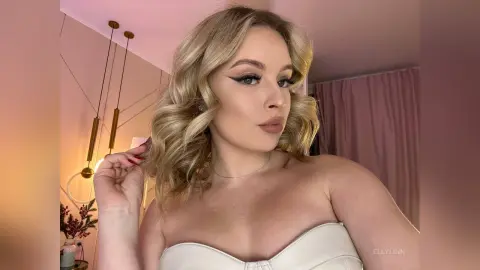 Blonde woman in a white strapless top posing with a soft, glamorous look in a bright indoor setting.