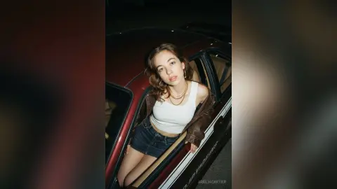 AmeliaCartier poses in a dark car wearing a white tank top, denim shorts, and a brown leather jacket, looking at the camera with brown hair.