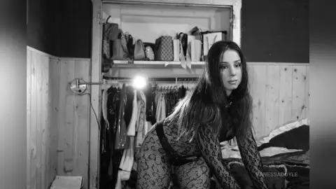 Moody black and white portrait of a woman in a patterned bodysuit sitting in a bedroom near a clothing rack.